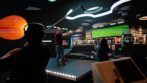 《og体育》的导演杰森·墨菲站在一个虚拟宇宙飞船的舞台上. 他正在和一名工作人员交谈，另一名工作人员拿着吊杆麦克风.